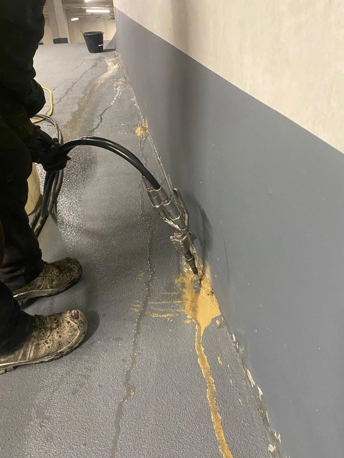 A worker operates a hose in a parking garage, applying a substance to a wall with visible leakage and grey flooring.